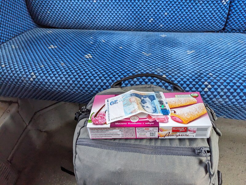 A box of cookies and a 20 lev note on top of a backpack on a train floor